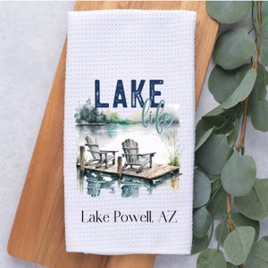 May include: White kitchen towel with a watercolor illustration of a lake scene with two chairs on a dock. The text "LAKE life" is in blue lettering above the scene. The text "Lake Powell, AZ" is in blue lettering below the scene.