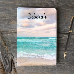 May include: A personalized notebook with a beach scene on the cover. The cover features a blue ocean with white waves crashing on a sandy beach. The sky is a light blue with white clouds. The name "Deborah" is printed in black cursive on the cover.
