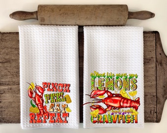 May include: Two white waffle weave kitchen towels with colorful crawfish and text designs. One towel reads "Pinch, Peel, Eat, Repeat" and the other says "When life gives you lemons, buy some crawfish."
