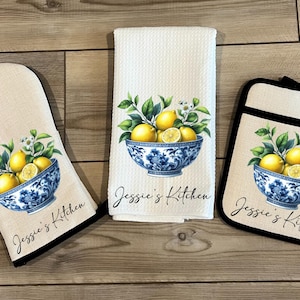 May include: A set of three kitchen items featuring a blue and white bowl filled with lemons and green leaves. The items include a tea towel, a pot holder, and an oven mitt. Each item has the text "Jessie's Kitchen" printed on it.