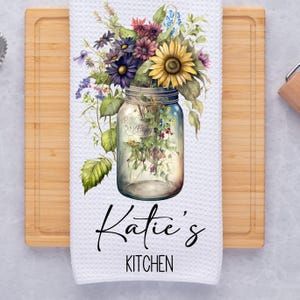 May include: White kitchen towel with a watercolor illustration of a mason jar filled with colorful wildflowers. The text "Katie's Kitchen" is printed below the jar.