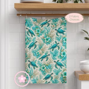 May include: A rectangular kitchen towel featuring a repeating pattern of teal sea turtles, light blue coral, and white plumeria flowers on a cream background. A circular pink badge in the corner displays the text 2 Fabric Choices.