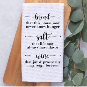 May include: White kitchen towel with a waffle weave texture. The towel features black script with the words "bread," "salt," and "wine," each followed by a short phrase. The towel is displayed on a wooden cutting board with greenery.