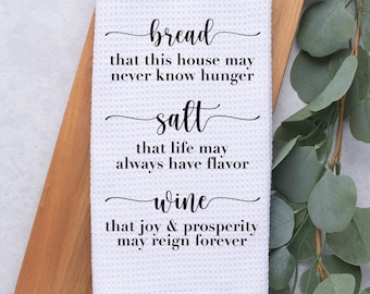 May include: A white waffle-weave kitchen towel featuring black text that reads: bread that this house may never know hunger, salt that life may always have flavor, and wine that joy & prosperity may reign forever.