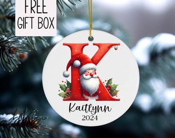 May include: A white ceramic Christmas ornament with a red letter "K" featuring Santa Claus. The ornament includes the name "Kaitlynn" and the year "2024". A gold string is attached for hanging. The words "FREE GIFT BOX" are in the upper left corner.