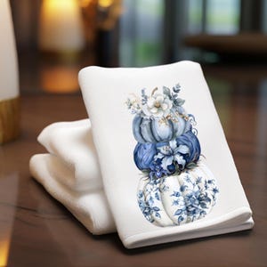 May include: White hand towel with a stack of three pumpkins in shades of blue and white. The pumpkins are decorated with floral designs and topped with white flowers and greenery. A stack of white towels is next to the decorated towel.