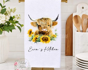 May include: A white waffle-weave kitchen towel featuring a printed illustration of a brown Highland cow surrounded by yellow sunflowers. The text "Erin's Kitchen" is printed in black script below the cow. The towel hangs on a gold metal stand.