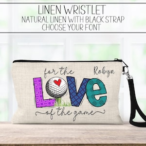 May include: A natural linen wristlet with a black strap. The wristlet features a golf ball with a heart on it and the words "for the love of the game" with the name "Robyn" above the golf ball.
