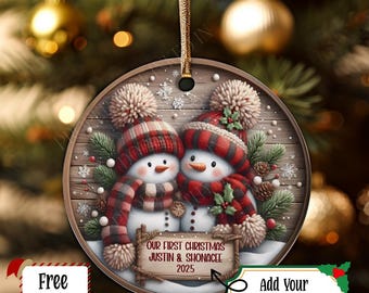 May include: Round Christmas ornament featuring two snowmen wearing red and black plaid hats and scarves. The ornament has a wooden background with the text "OUR FIRST CHRISTMAS JUSTIN & SHONACEE 2025". A gold ribbon is attached for hanging.
