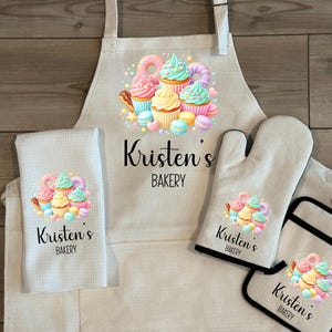 May include: A set of kitchen textiles featuring a cupcake design. Includes an apron, oven mitt, pot holder, and a kitchen towel. The text "Kristen's Bakery" is printed on each item. The image also includes the text "Available as a set or individually."