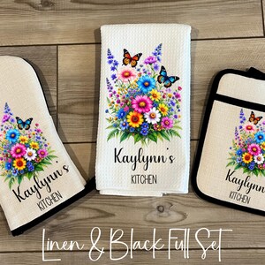 May include: A kitchen set featuring a beige oven mitt, a white kitchen towel, and a beige pot holder. Each item has a floral design with butterflies and the text "Kaylynn's Kitchen". The set is trimmed in black.