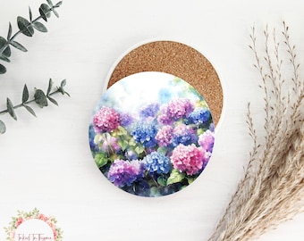 May include: A set of two round ceramic coasters featuring a watercolor painting of pink and blue hydrangeas. The coasters have a cork backing.