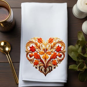 May include: White decorative towel featuring a heart-shaped design with autumn leaves in shades of red, orange, and gold. The leaves are intricately detailed with a gold filigree border. The towel is folded and placed on a dark wooden surface.
