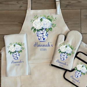 May include: A kitchen set featuring an apron, oven mitt, pot holder, and a towel. Each item has a floral design of white hydrangeas in a blue and white vase. The text "Hannah's Kitchen" is printed on each item. The text "Available as a set or individually" is at the top.