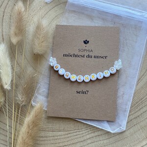 Könnte beinhalten: Eine weiße Karte mit dem Text "Sophia, möchtest du unser Blumenkind sein?" und ein weißes Perlenarmband mit goldenen Buchstaben, die "Blumenkind" buchstabieren.