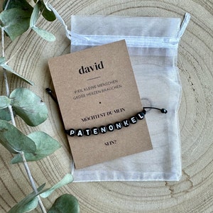 Könnte beinhalten: Ein schwarzes Perlenarmband mit der Aufschrift "PATENONKEL" wird auf einer Kraftpapierkarte mit dem Text "david" und deutschem Text angezeigt. Die Karte liegt in der Nähe eines durchsichtigen weißen Kordelzugsacks und Eukalyptusblättern.