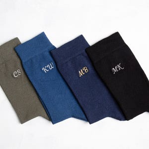 Puede incluir: Cuatro pares de calcetines de diferentes colores, cada uno con un monograma bordado diferente. Los calcetines son verdes, azules, azul marino y negros.