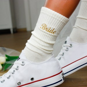 May include: Off-white ribbed crew socks featuring the word Bride embroidered in gold thread, paired with white low-top canvas sneakers that have silver eyelets and red and blue stripes along the rubber sole edge.