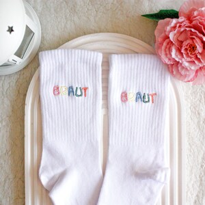 May include: A pair of white socks with the word "BRAUT" embroidered on each sock in rainbow colors.