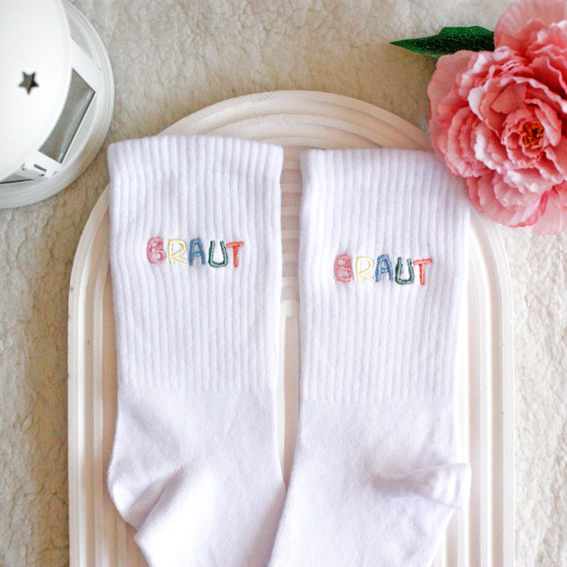 May include: A pair of white socks with the word "BRAUT" embroidered on each sock in rainbow colors.