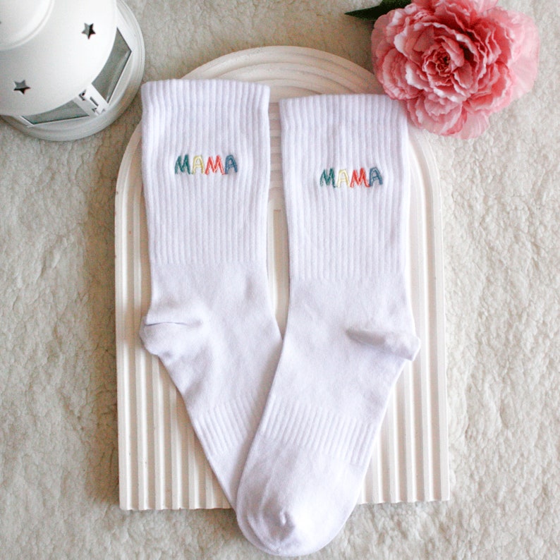 May include: A pair of white socks with the word "MAMA" embroidered on each sock in rainbow colors.