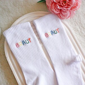 May include: A pair of white socks with the word "BRAUT" embroidered in rainbow colors.