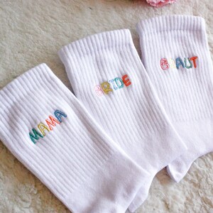 May include: Three white socks with colorful embroidered text. The socks say "Mama", "Bride", and "Braut".