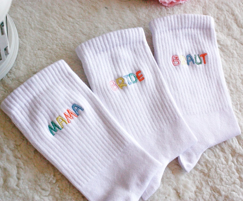 May include: Three white socks with colorful embroidered text. The socks say "Mama", "Bride", and "Braut".