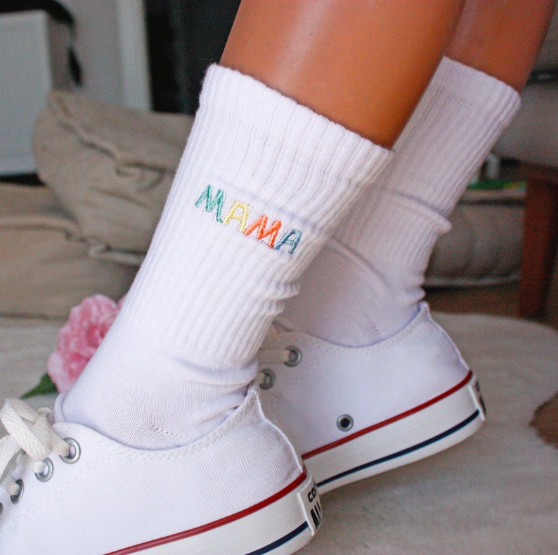May include: White socks with the word "MAMA" embroidered in rainbow colors. The socks are worn with white sneakers.