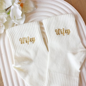 May include: A pair of white ankle socks with gold embroidery that says "Wifey".
