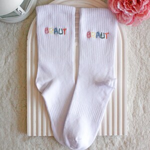 May include: A pair of white socks with the word "BRAUT" embroidered on each sock in rainbow colors.