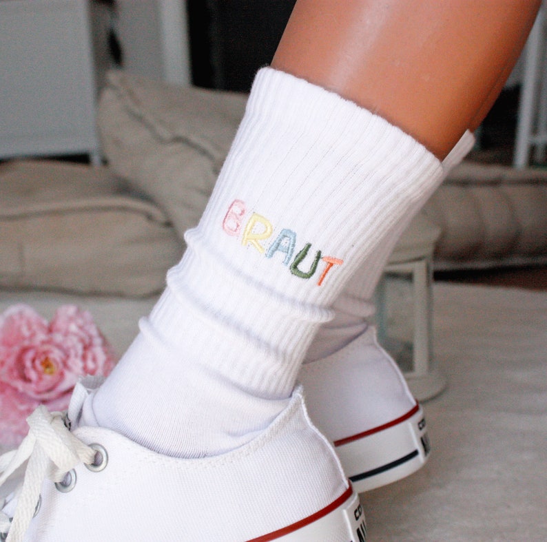 May include: White knee-high socks with the word "BRAUT" embroidered in rainbow colors. The socks are worn with white sneakers.