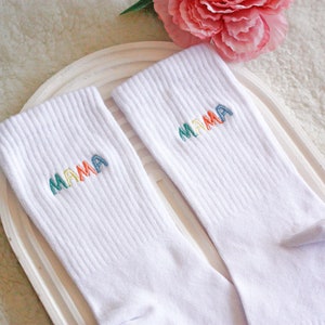 May include: A pair of white socks with the word "MAMA" embroidered on each sock in rainbow colors.
