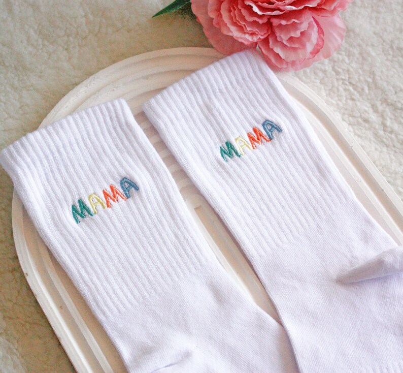 May include: A pair of white socks with the word "MAMA" embroidered on each sock in rainbow colors.