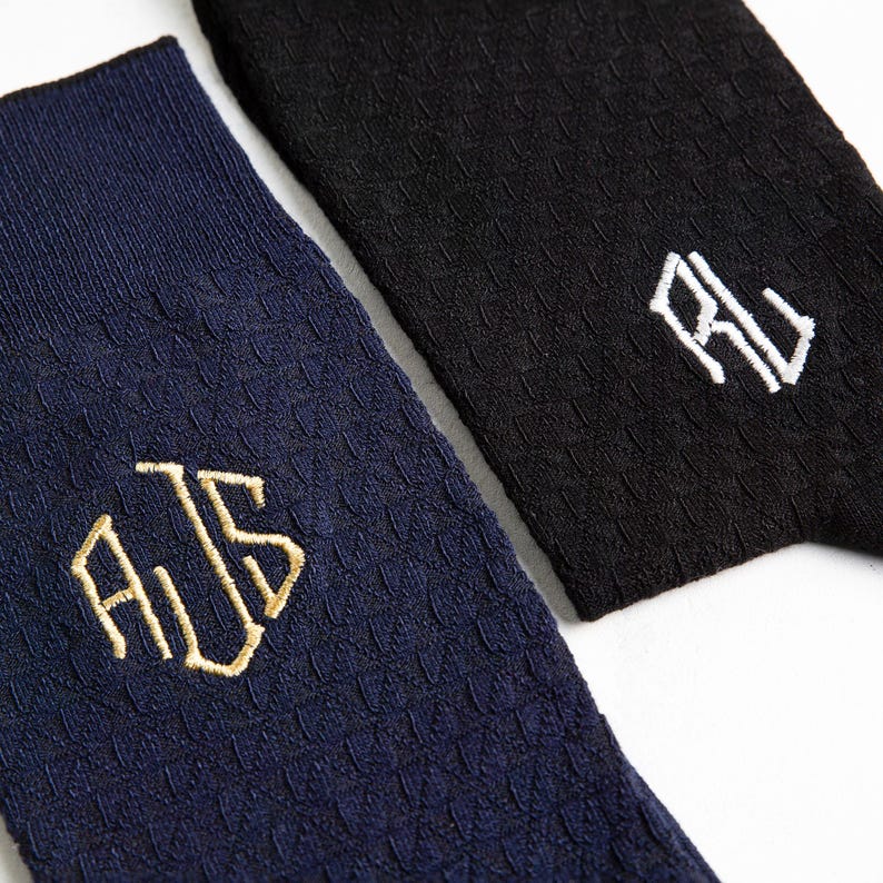Puede incluir: Dos pares de calcetines con iniciales bordadas. Un par es azul marino con iniciales doradas "AJS" y el otro par es negro con iniciales blancas "RJ".