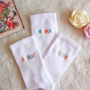 May include: Three white socks with colorful embroidered words. The socks say "BRIDE", "MAMA", and "BRAUT".