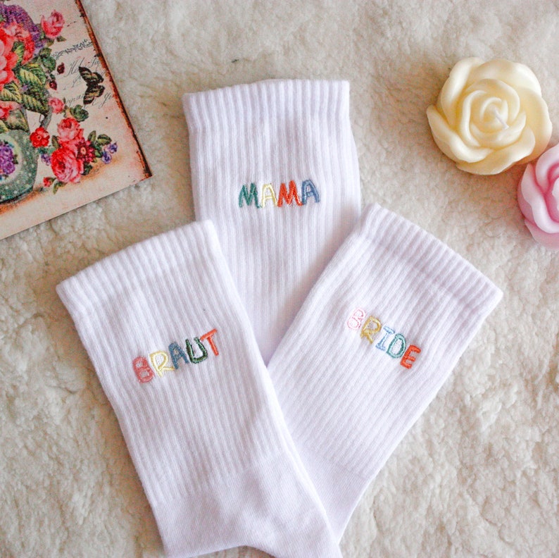 May include: Three white socks with colorful embroidered words. The socks say "BRIDE", "MAMA", and "BRAUT".