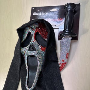 Può includere: Un set di costumi di Halloween con una maschera Ghost Face tempestata di strass con cappuccio nero e un coltello decorativo. La maschera ha strass iridescenti e accenti rossi. Il coltello è anch'esso impreziosito da strass e vernice rossa.