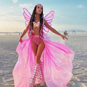 Puede incluir: Una mujer con un disfraz de mariposa rosa con alas iridiscentes, un sujetador de mariposa rosa y una falda rosa. Lleva botas de plataforma rosa con m&uacute;ltiples hebillas. La mujer est&aacute; de pie en una playa de arena blanca.