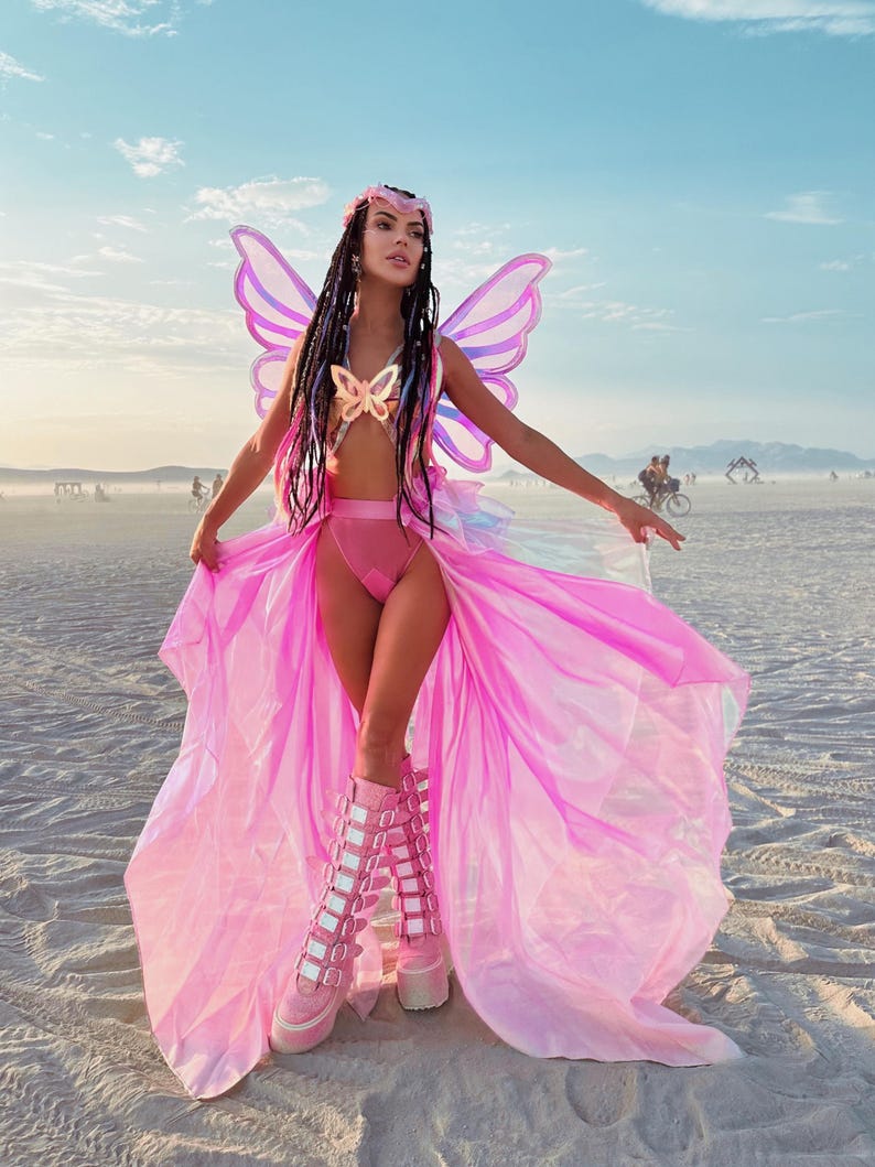 Puede incluir: Una mujer con un disfraz de mariposa rosa con alas iridiscentes, un sujetador de mariposa rosa y una falda rosa. Lleva botas de plataforma rosa con m&uacute;ltiples hebillas. La mujer est&aacute; de pie en una playa de arena blanca.