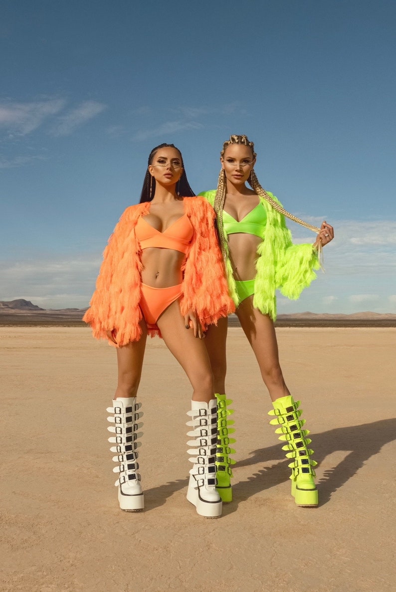 Pu&ograve; includere: Due donne con abiti color neon e stivali platform alti con fibbie. La donna a sinistra indossa una giacca di pelliccia arancione brillante e un top e un fondo bikini abbinati. La donna a destra indossa una giacca di pelliccia verde brillante e un top e un fondo bikini abbinati. Entrambe le donne indossano stivali platform bianchi con fibbie nere.