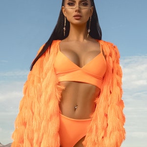 Pu&ograve; includere: Una donna indossa una giacca arancione brillante con frange e un top e un fondo bikini abbinati. &Egrave; in piedi in un paesaggio desertico con un cielo blu sullo sfondo.