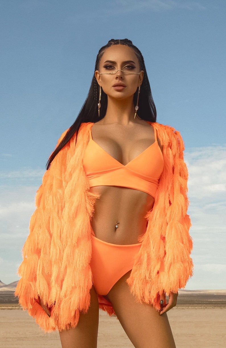 Pu&ograve; includere: Una donna indossa una giacca arancione brillante con frange e un top e un fondo bikini abbinati. &Egrave; in piedi in un paesaggio desertico con un cielo blu sullo sfondo.