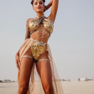 May include: A gold and tan two-piece outfit with a halter top and high-waisted bottoms. The outfit has a leopard print design and is accessorized with gold chains and leg bands. A sheer, tan skirt drapes from the waist.