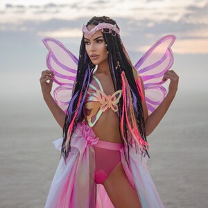Puede incluir: Una mujer con un disfraz de mariposa rosa con alas iridiscentes, un top con forma de mariposa y una falda rosa. Tiene trenzas largas y coloridas y una corona dorada.