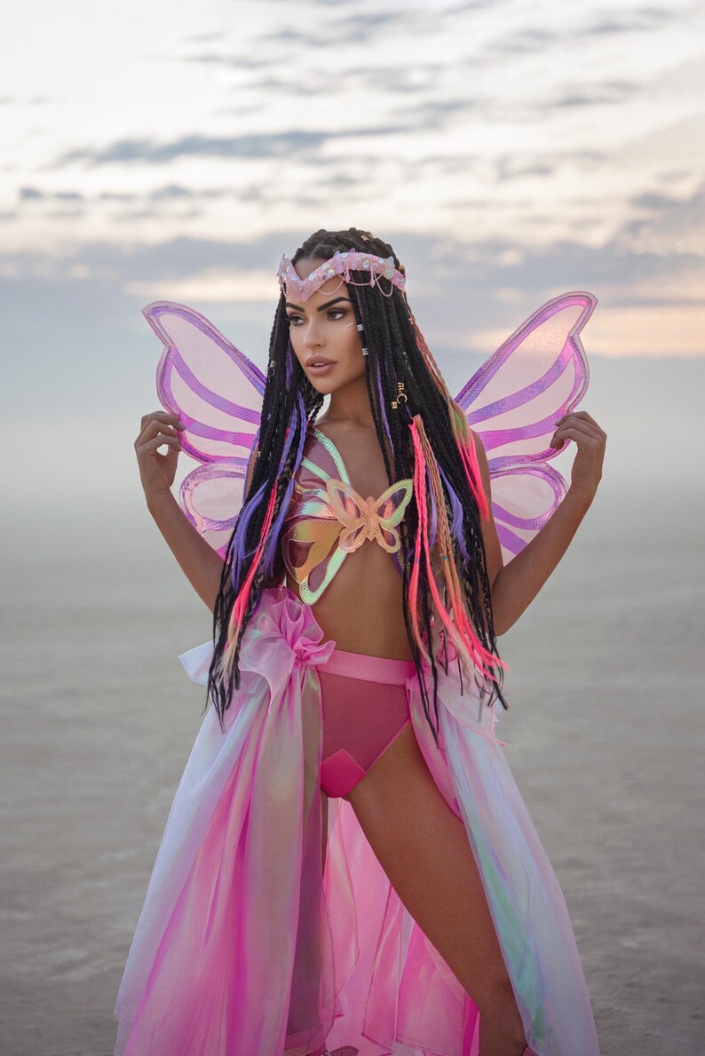 Puede incluir: Una mujer con un disfraz de mariposa rosa con alas iridiscentes, un top con forma de mariposa y una falda rosa. Tiene trenzas largas y coloridas y una corona dorada.