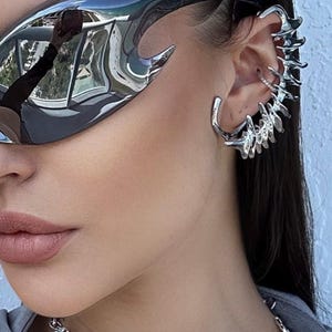 May include: A silver chain necklace with flame-shaped links. The necklace is worn by a person with dark hair and sunglasses. The person is also wearing a silver ear cuff with multiple spikes.