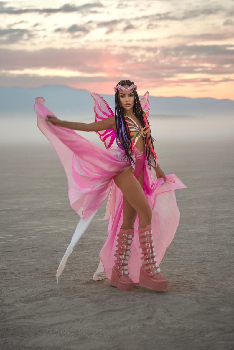 Puede incluir: Una mujer con un disfraz de mariposa rosa con un top de mariposa dorado y botas de plataforma rosa. El disfraz tiene una falda larga y fluida y alas de mariposa. La mujer est&aacute; de pie en un entorno des&eacute;rtico con un cielo rosa en el fondo.