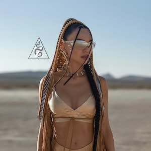 May include: A gold bikini top and matching bottoms with a snakeskin pattern. The model is wearing silver-framed sunglasses and a spiked choker. Long braids with fringe and a logo with a snake are also visible.