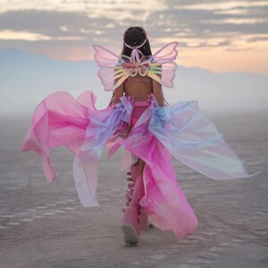 Puede incluir: Una persona vestida con un disfraz de mariposa rosa e iridiscente con alas y una falda fluida, caminando sobre una superficie blanca y polvorienta.